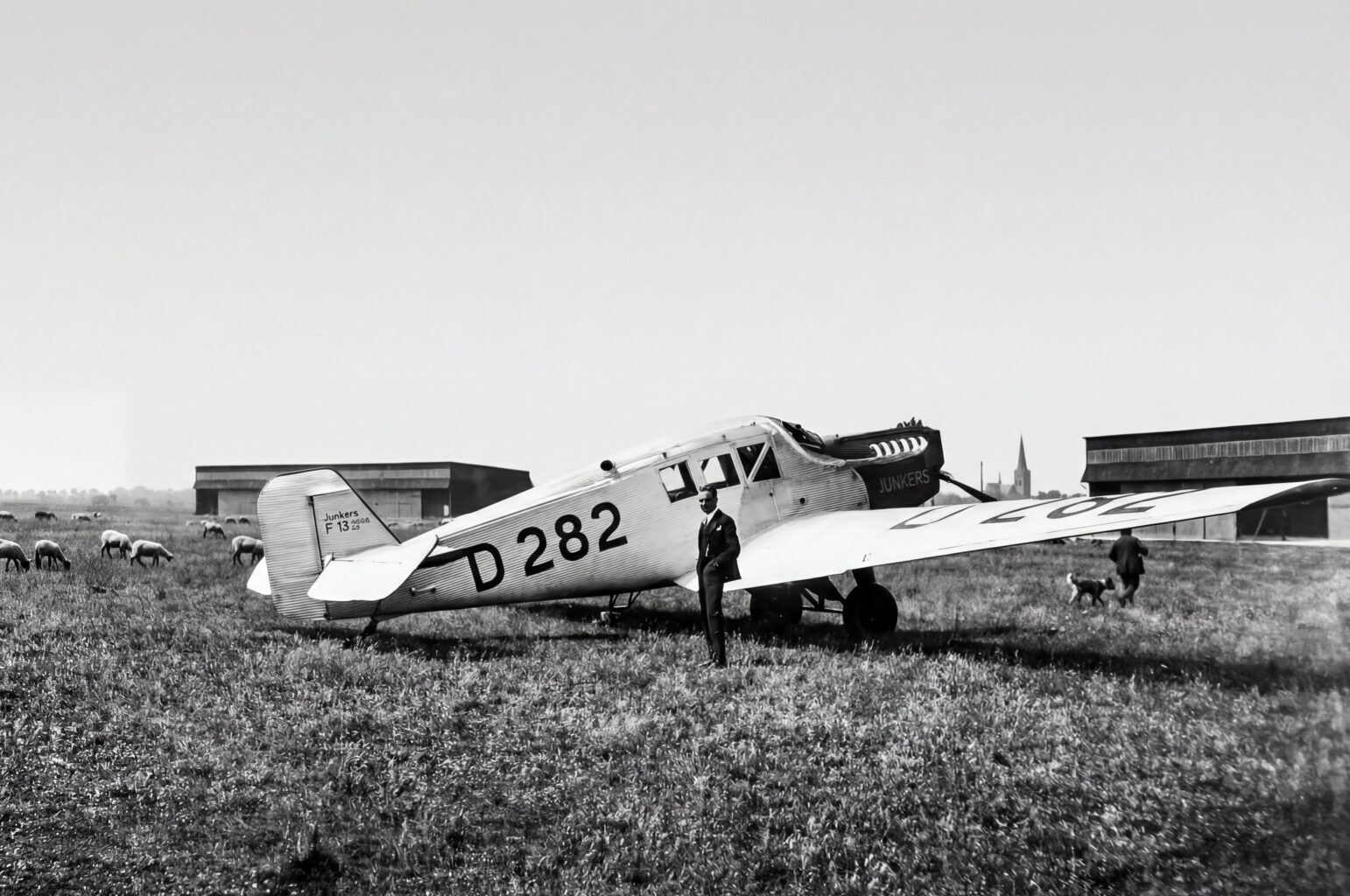 Flugzeug vor Schafherde: Die Junkers F13 auf dem Flugplatz Merzbrück. Die Konstruktion aus dem Jahr 1919 war nicht nur das erste Ganzmetallflugzeug der zivilen Luftfahrt, sondern auch das Arbeitstier der frühen Lufthansa. Ein knappes Drittel der Flugzeugflotte von 162 Maschinen im Gründungsjahr 1926 entfällt auf diesen Flugzeugtyp, insgesamt 47 Maschinen. (Foto: Archiv Forschungsflugplatz Würselen-Aachen GmbH, SW-Original und Farbversion des Bildes KI-bearbeitet)