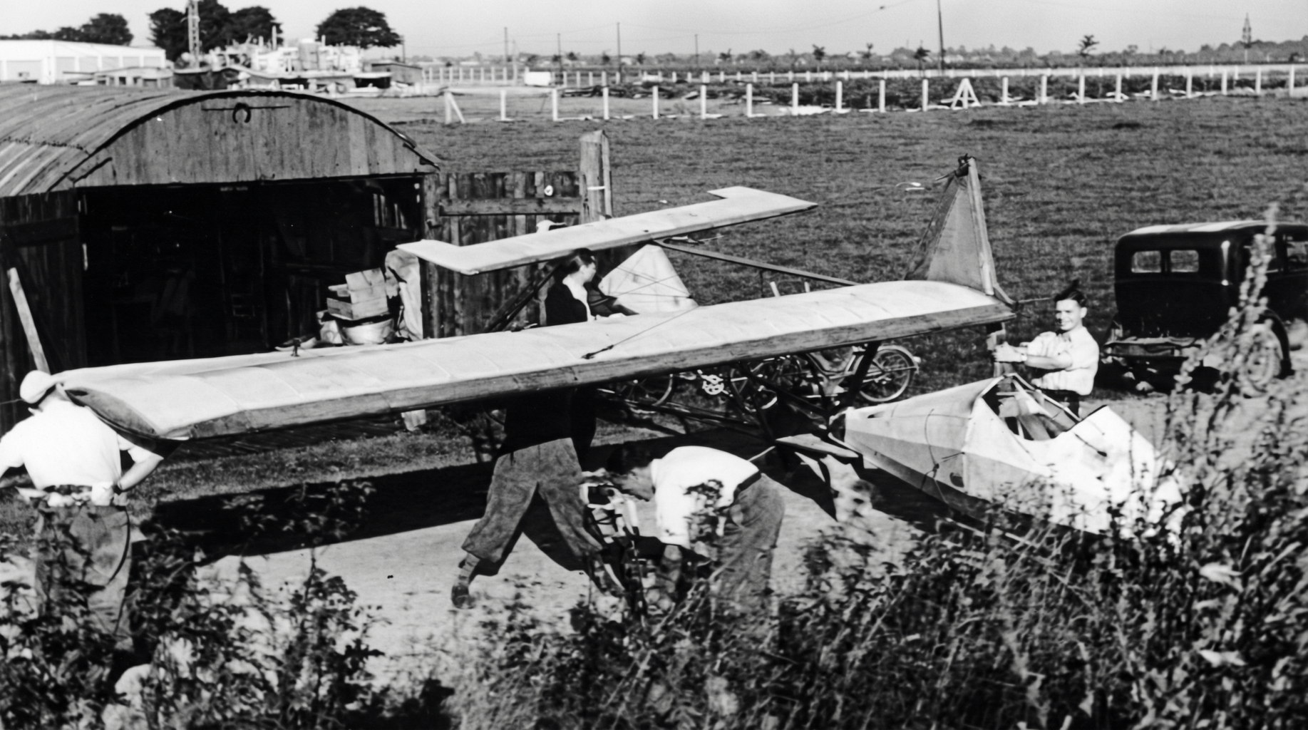 Die Nissenhütte ist zu klein, um Flugzeuge aufgerüstet hinein zu schieben. Deshalb muss auch der Schulgleiter SG38 vor jedem Flugbetrieb aufgerüstet werden – ein mühsames Geschäft, müssen Tragflügel und Leitwerk doch aufwendig mit Drähten verspannt werden. Die korrekte Spannung wird von erfahrenen Vereinsmitgliedern durch Klangtest ermittelt: Sie wird wie eine Geigenseite gezupft ( Foto: Luftsportverein Aachen e.V. SW-Original und Farbversion des Bildes KI-bearbeitet)